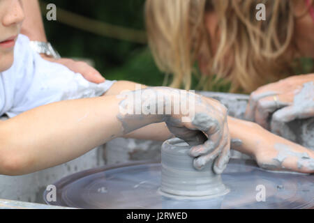 Atelier di ceramica, di insegnamento in ceramica. Una tazza di argilla è essendo formata da un giovane bambino su un tornio del vasaio con insegnante in background. Foto Stock