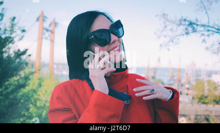 Giovane donna in rosso cappotto e occhiali da sole a parlare sul suo smartphone e sorridente Foto Stock