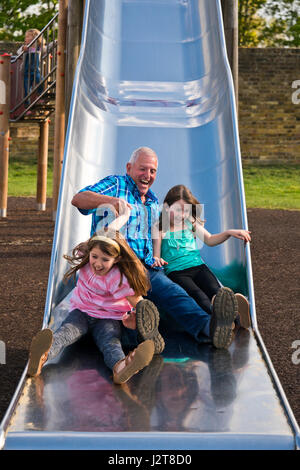 Ritratto verticale di un nonno lo scorrimento verso il basso una diapositiva con i suoi nipoti in un parco giochi. Foto Stock