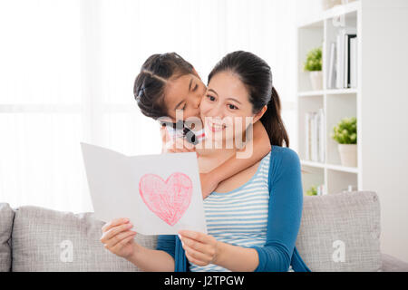 Carino piccola figlia bacio della donna guancia quando la madre ha ricevuto l'amore dono della scheda e leggere tranquillamente seduto sul divano del soggiorno in Foto Stock