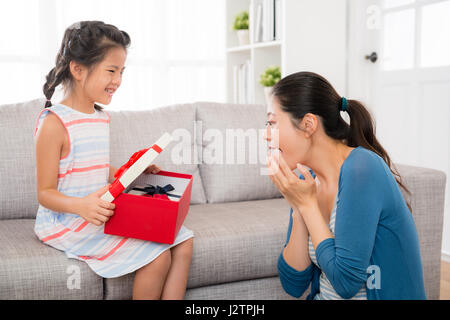 Sorpreso madre felice di guardare la apertura della giornata della madre confezione regalo dalla graziosa piccola figlia amd insieme nel salotto di casa shari Foto Stock