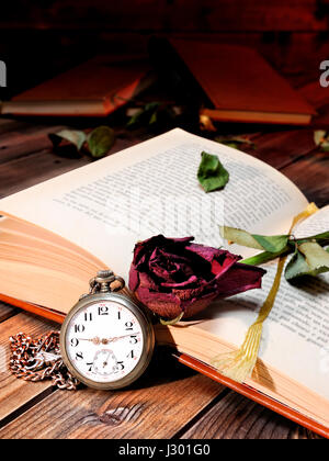 Una red rose appassite attraverso le pagine di un vecchio libro ingiallito dal tempo e un antico orologio da tasca su un vecchio tavolo di legno. Foto Stock