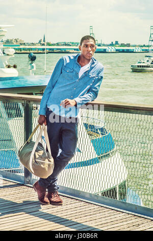 In viaggio. Indossando camicia a maniche lunghe, pantaloni blu, marrone boot scarpe, una mano portando un borsone, un giovane nero studente di college è in piedi sul dock da Foto Stock