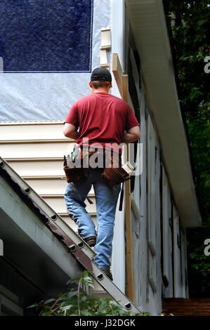 Proprietario fa un auto rata lavoro sulla sua casa schierati. Egli è in piedi su una scala attaccata al tetto della sua casa. Foto Stock