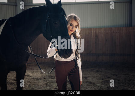 Bellissima ed elegante ragazza bionda sta in piedi vicino al suo cavallo nero medicazione di concorrenza uniforme camicetta bianca maglietta e pantaloni marrone. Piscina portraite in Foto Stock