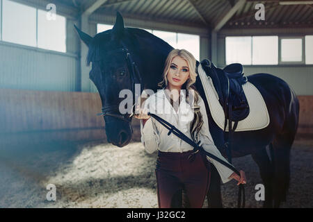 Bellissima ed elegante ragazza bionda sta in piedi vicino al suo cavallo nero medicazione di concorrenza uniforme camicetta bianca maglietta e pantaloni marrone. Piscina portraite in Foto Stock