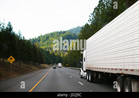 Semi-camion Classic Big RIG con rimorchio frigorifero su una tortuosa strada di montagna che passa nel mezzo di alberi verdi che lo circondano e l'insegna dell'autostrada Foto Stock