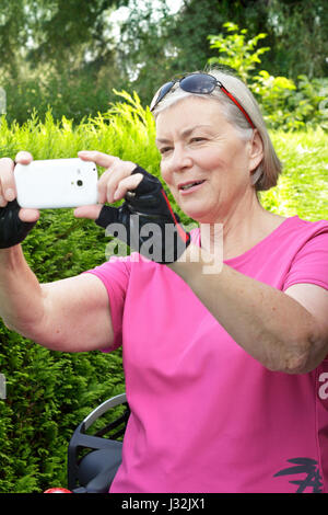 Donna senior all'aperto in estate con il rosa sport shirt, ciclismo guanti e occhiali da sole di scattare una foto con il suo smartphone, sfondo verde Foto Stock