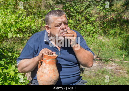 Ucraino contadino senior prendere un vecchio brocca di argilla e il fumo di sigaretta da laminati di sé con un vecchio giornale di rottami e di tabacco coltivate su un proprio g Foto Stock