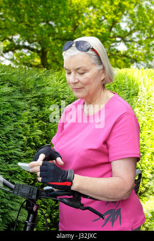 Donna senior all'aperto in estate con il rosa sport shirt, ciclismo guanti e occhiali da sole guardando il suo smartphone, siepe verde in background Foto Stock