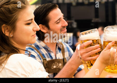 Coppia giovane in tradizionali bavaresi Tracht - Dirndl e lederhosen - in una tenda di birra al Oktoberfest a Monaco di Baviera la tostatura con loro 1 litro boccale di birra Foto Stock
