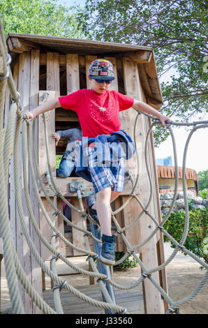 Un giovane ragazzo attraversa un ponte di corde che è parte di un telaio di arrampicata in un parco giochi per bambini. Foto Stock