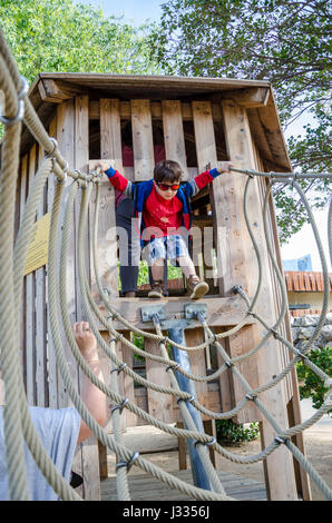 Un giovane ragazzo attraversa un ponte di corde che è parte di un telaio di arrampicata in un parco giochi per bambini. Foto Stock