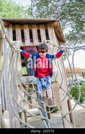 Un giovane ragazzo attraversa un ponte di corde che è parte di un telaio di arrampicata in un parco giochi per bambini. Foto Stock