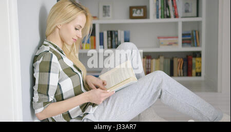 Vista laterale del giovane ragazza in abbigliamento casa seduti sul pavimento con libro su sfondo di scaffali. Foto Stock