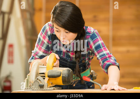 Carpenter utilizzando una sega circolare. femmina falegname azienda tavola circolare sega elettrica il taglio di legno in officina. razza mista asiatica modello cinese Foto Stock