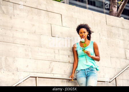 Ragazza in attesa per voi. Tenendo la rosa bianca, indossando il serbatoio verde top, jeans, orecchini a pendente, capelli fluttuante nel vento, giovani African American woman standing b Foto Stock