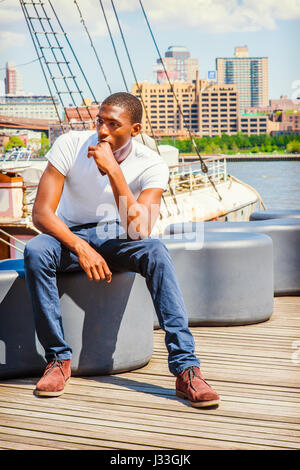 Giovane americano africano uomo che viaggia in New York, indossando il bianco di T-shirt, pantaloni blu, marrone boot scarpe, seduto sul ponte sul fiume, mano toccando la bocca, Foto Stock