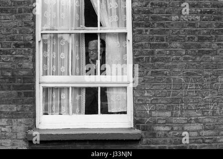 Da solo, solitario, un anziano anziano che guarda fuori dalla sua finestra attraverso le tende a rete, 1970s Whitechapel, East End Londra 1975 UK HOMER SYKES Foto Stock