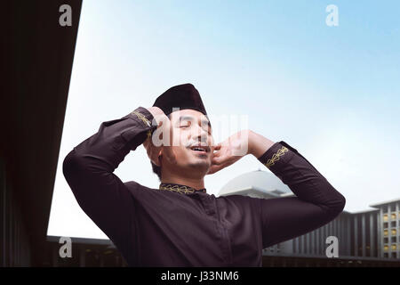 Bello musulmani asiatici uomo in abito tradizionale pregando con sfondo moschea Foto Stock