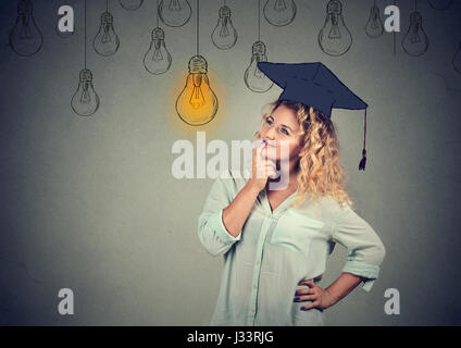 Considerato studente laureato ragazza giovane donna nel cappuccio camice cercando fino alla luce brillante lampadina pensando isolato muro grigio Sfondo. Cerimonia di laurea f Foto Stock