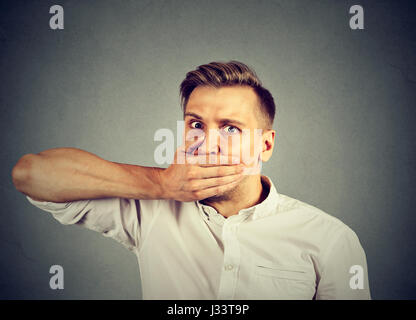 Spaventata giovane uomo che copre la bocca con la mano isolato sul muro grigio Sfondo. Emozioni umane espressione faccia Foto Stock