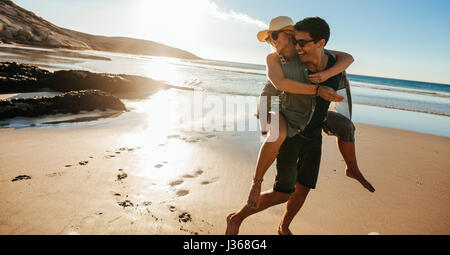 L uomo dando piggyback ride alla ragazza sulla spiaggia. Felice coppia giovane divertirsi in riva al mare, godendo le vacanze estive. Foto Stock