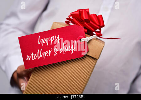Primo piano di un giovane uomo caucasico in una camicia bianca tenendo una confezione regalo con un etichetta rossa legata ad essa con il testo felice festa della mamma scritto in esso Foto Stock