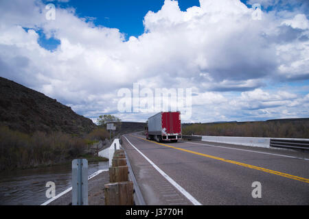 Semi in movimento il carrello con il carico in un dry van rimorchio con porte rosso sulla divisa corsie passare oltre il ponte su un piccolo fiume che attraversa Foto Stock