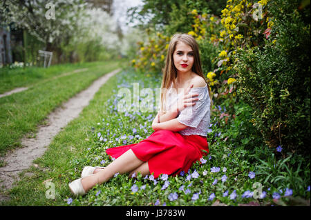 Ritratto di sitiing bellissima ragazza con labbra rosse a primavera sbocciano i fiori del giardino di erba con fiori, indossare il vestito rosso e la camicia bianca. Foto Stock