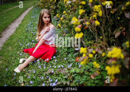 Ritratto di sitiing bellissima ragazza con labbra rosse a primavera sbocciano i fiori del giardino di erba con fiori, indossare il vestito rosso e la camicia bianca. Foto Stock