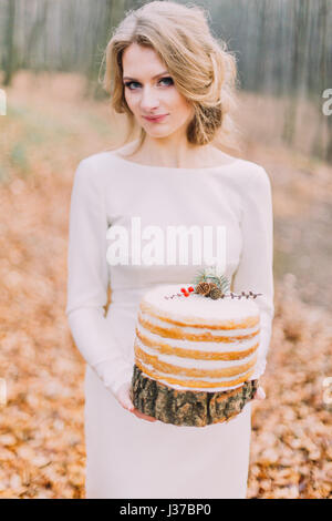 Attraente bionda sposa con torta di nozze nella foresta di autunno Foto Stock