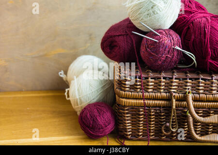 Vintage cesto in vimini sfere bugne di rosso bianco filati di lana, aghi sulla tavola di legno, lavorazione a maglia, artigianato, hobby concetto, close up Foto Stock