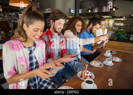 Sorridendo gli amici usando i loro cellulari durante la seduta di tazze da caffè e dessert sul tavolo nel ristorante Foto Stock