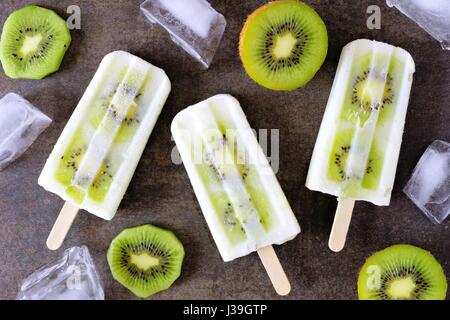 Kiwi yogurt alla vaniglia popsicles con ghiaccio e frutta tagliata su una pietra scura sullo sfondo Foto Stock