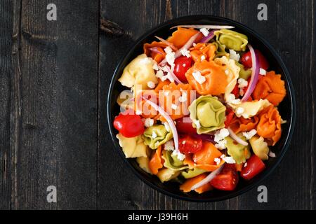 Ciotola multicolore di tortellini con insalata di pomodoro e cipolle, vista aerea su un legno scuro dello sfondo Foto Stock