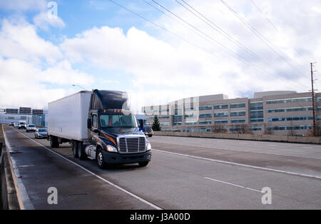 Moderna attrezzatura scuro cabina giorno semi carrello con spoiler sulla parte superiore con un secco van rimorchio è in movimento nella parte anteriore della colonna di trasporto da una ampia strada multilane Foto Stock