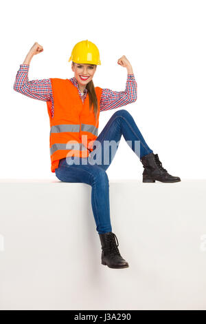 Sorridente giovane donna in giallo hardhat, arancione giubbotto lumberjack, t-shirt, jeans e stivali neri, seduto sulla cima di una casella bianca, flettendo i muscoli un Foto Stock