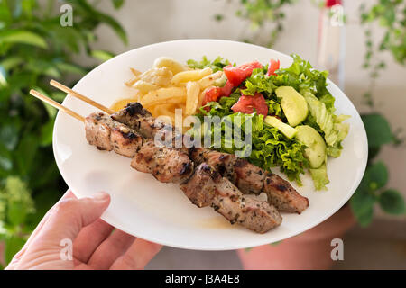 Una piastra di souvlaki di maiale con patate fritte e insalata verde con pomodori, lattuga e cetriolo, home background. Foto Stock