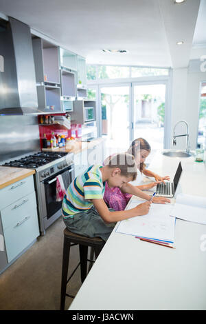 Ragazzo che sta facendo il suo dovere mentre ragazza utilizzando laptop in cucina a casa Foto Stock