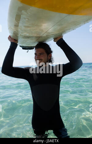 Surfista sorridente in portante muta surfboard sopra la testa al litorale Foto Stock