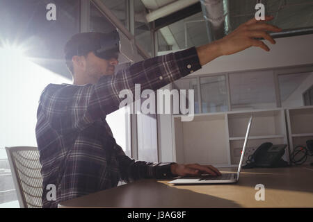 Vista laterale di imprenditore che indossa la realtà virtuale cuffie mentre utilizzando computer portatile in ufficio Foto Stock