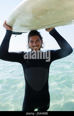 Ritratto di sorridere surfer in portante muta surfboard sopra la testa al litorale Foto Stock