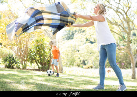 Madre di diffondere la coperta picnic mentre il figlio a giocare a calcio in background in una giornata di sole Foto Stock