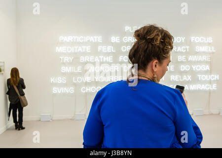 New York, US. 05 Maggio, 2017. New York, NY - 5 maggio 2017. Il giorno di apertura del fregio Art Fair, presentazione di arte moderna e contemporanea presentata da gallerie provenienti da tutto il mondo, su Randall's Island in New York City. Una donna controlla il suo telefono davanti a un'installazione di tubi al neon da Jeppe Hein, "Si prega di partecipare,' in 303 Gallery. Credit: Ed Lefkowicz/Alamy Live News Foto Stock