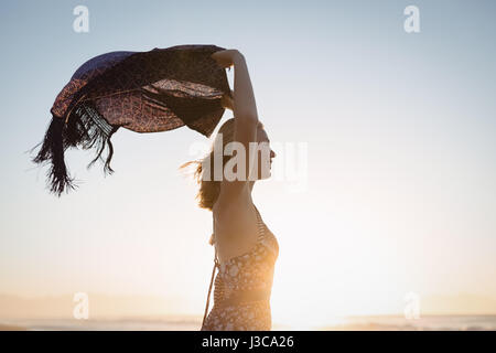 Vista laterale della donna felice azienda sciarpa contro il cielo chiaro in spiaggia durante il tramonto Foto Stock