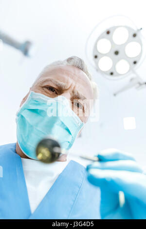Professional dentista maturo nella maschera di medico tenendo la bocca a specchio e guardando la fotocamera Foto Stock