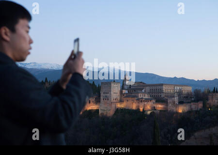 Asian tourist usa il suo smartphone per fotografare il Palazzo Alhambra dal Mirador de San Nicolas in El Albaycin distretto in Granada, Andalusia, Spagna. Il Nasrid palazzi con la Torre de Comares, la chiesa di Santa Maria de la Alhambra e il Palazzo di Carlo V sono visibili nella foto da sinistra a destra. Foto Stock