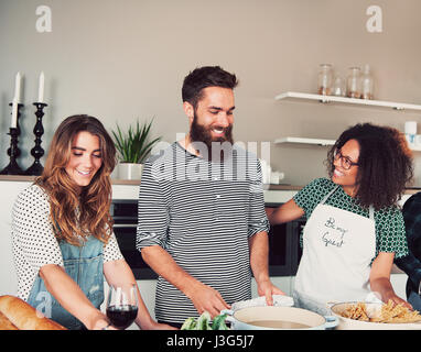 Tre felice giovane adulto maschio diversificato e amici avente una parte di cottura all'interno. Pane e gli ingredienti sul tavolo di fronte a loro. Foto Stock