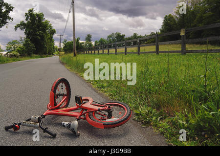 Rosso del bambino giacente in bicicletta su una strada rurale rappresenta i pericoli dei bambini equitazione biciclette senza caschi Foto Stock